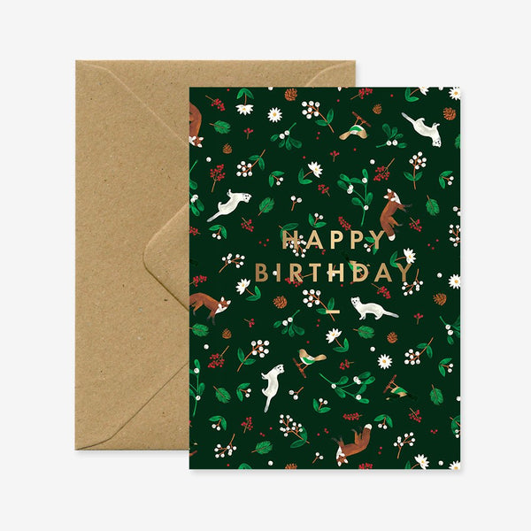 A birthday greeting card with a green floral pattern and gold foil details, displaying the text 'Happy Birthday'. It is accompanied by a brown envelope.