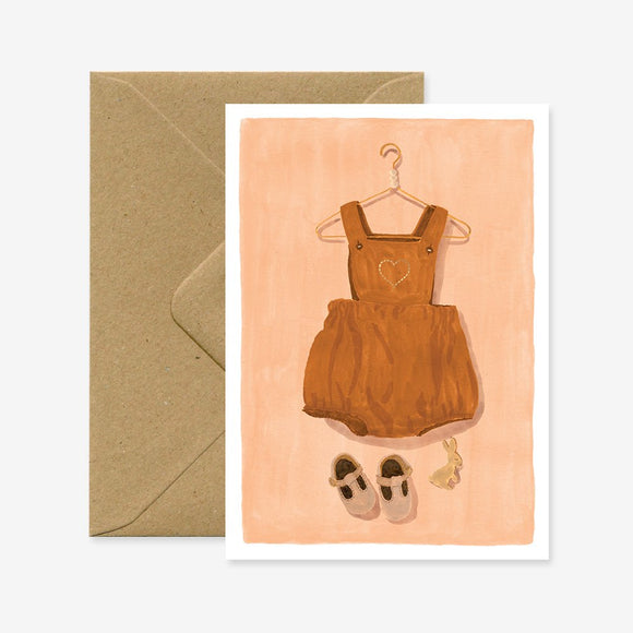 A baby greeting card with a illustration of a brown baby romper suit and shoes on a pink background, accompanied by a brown envelope.