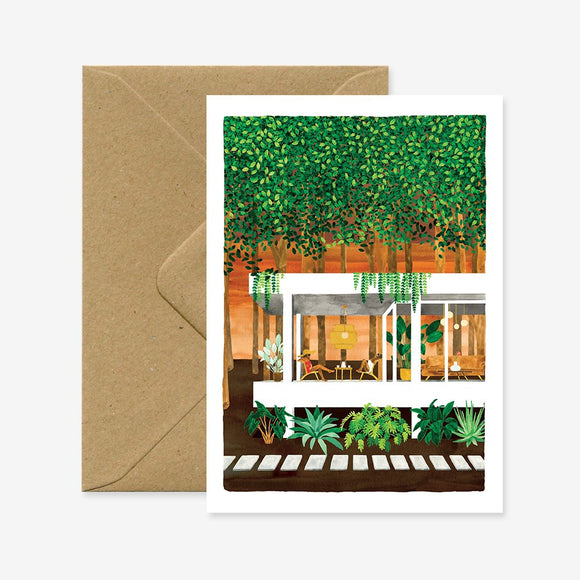 A greeting card with an illustration of a house in the woods, accompanied by a brown envelope.