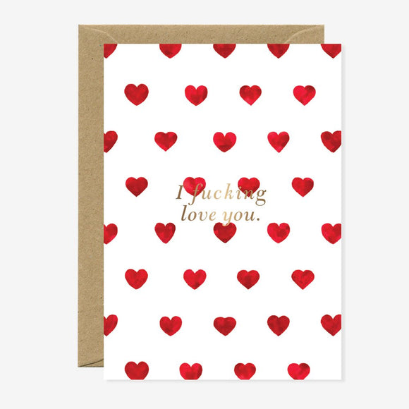 A white greeting card with a pattern of red hearts and the phrase 'I f*cking love you' in gold foil at the center. A brown envelope is included.