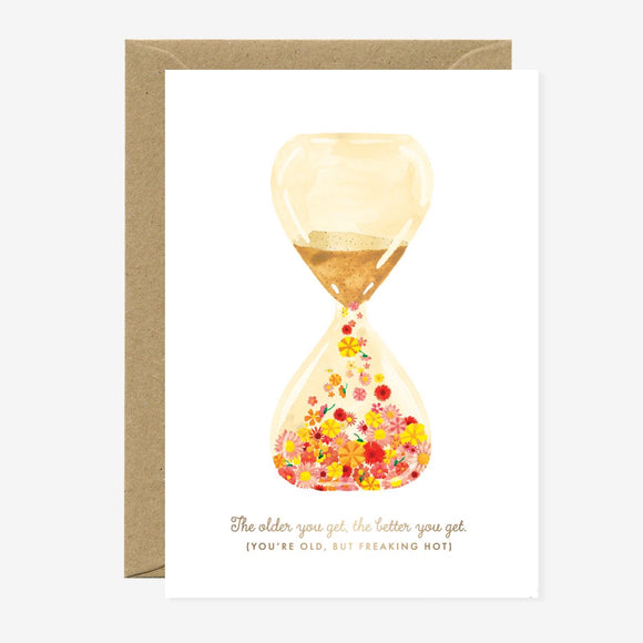 A birthday greeting card featuring a graphic of an hourglass with gold foil details and multicolored sand, alongside a brown envelope.