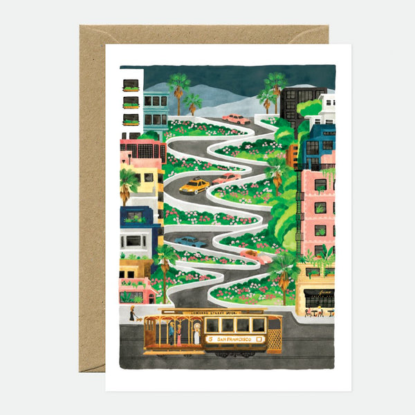 A greeting card featuring an illustration of a winding road with houses and a cable car, suggestive of a travel landscape. The card is accompanied by a brown envelope.