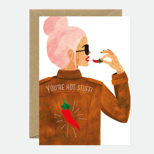 A greeting card with an illustration of a woman with pink hair and sunglasses, wearing a brown jacket with a chili pepper design, with the text 'YOU'RE HOT STUFF!' The card is accompanied by a brown envelope.