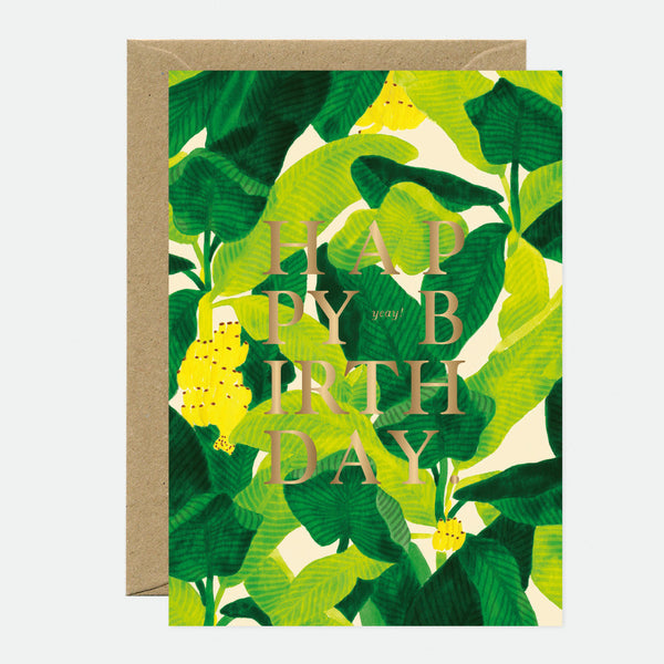 A birthday greeting card with gold foil text that reads 'HAPPY BIRTHDAY', featuring a green leafy background, accompanied by a brown envelope.