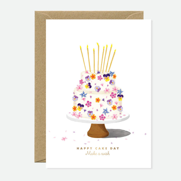 A greeting card featuring a gold foil illustration of a flower cake with candles on it, and the text 'Happy Cake Day' written at the bottom. A brown envelope is included.