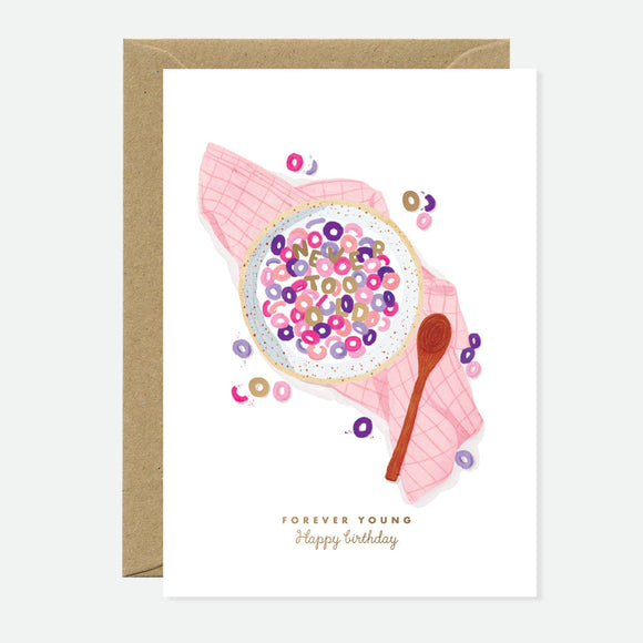A greeting card with a illustration of a bowl of cereal with the words 'FOREVER YOUNG' written across it, accompanied by a spoon and scattered cereal pieces. The card is placed against a beige background.