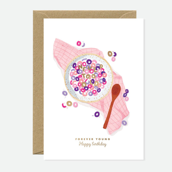 A greeting card with a illustration of a bowl of cereal with the words 'FOREVER YOUNG' written across it, accompanied by a spoon and scattered cereal pieces. The card is placed against a beige background.