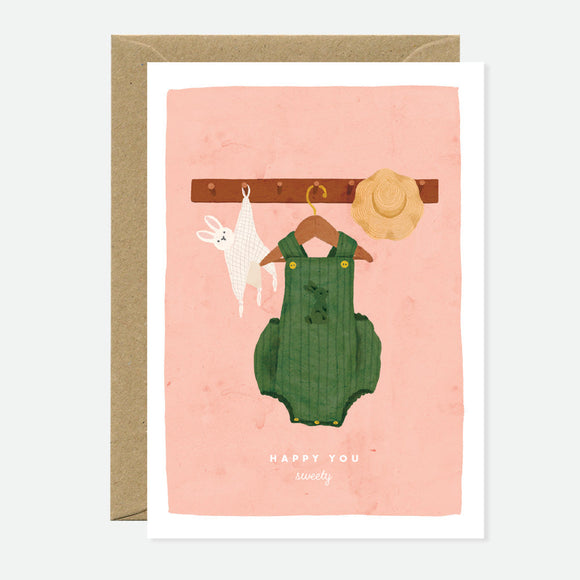A greeting card with an illustration of a rabbit in green overalls hanging on a clothesline, with the text 'HAPPY YOU' on a pink background. A brown envelope accompanies the card.