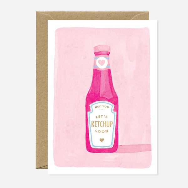 A greeting card with an illustration of a ketchup bottle on a pink background, with the text 'let's KETCHUP soon' on it. There is a brown envelope behind the card.