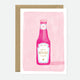 A greeting card with an illustration of a ketchup bottle on a pink background, with the text 'let's KETCHUP soon' on it. There is a brown envelope behind the card.