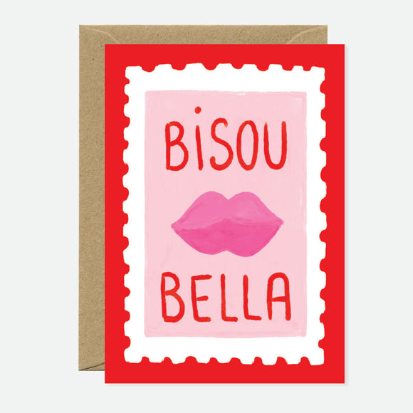 A greeting card with a pink background and red border featuring the text 'BISOU BELLA' with a pair of pink lips illustration above the text. The card is accompanied by a brown envelope.