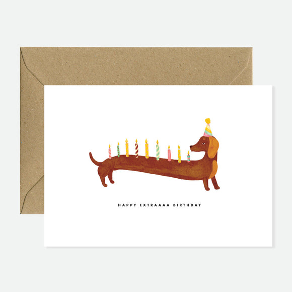 A greeting card with an illustration of a dachshund wearing a party hat with candles, indicating it is for a birthday. The card is displayed with a natural white envelope.