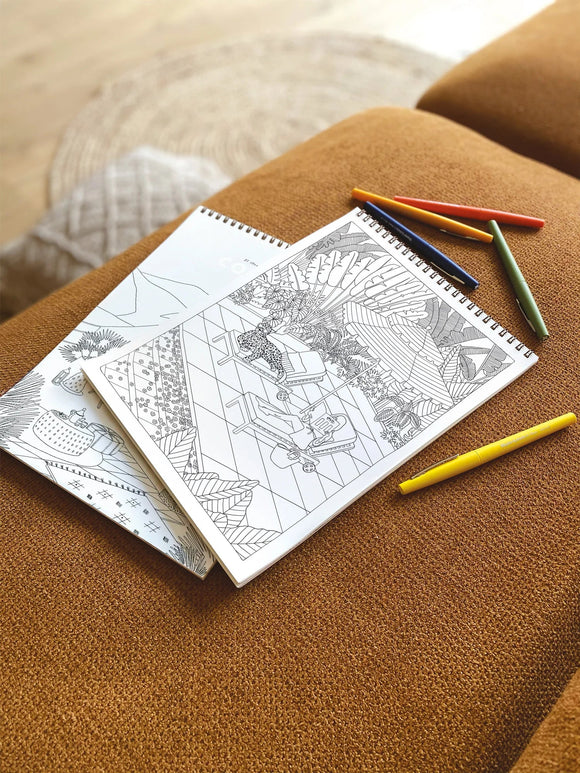 Open spiral coloring book with botanical illustration and markers placed on a brown fabric surface