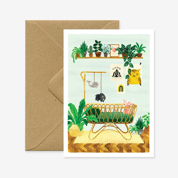 A baby greeting card with a illustration of a baby in a crib with decorative elements like plants, a teddy bear, and a mobile. Accompanied by a brown envelope.