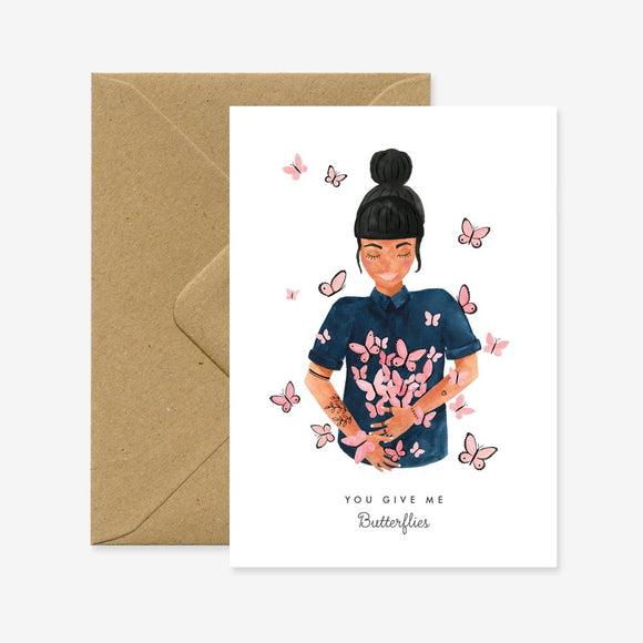 A greeting card with an illustration of a woman surrounded by butterflies, with a brown envelope in the background. The text on the card reads 'You give me Butterflies.