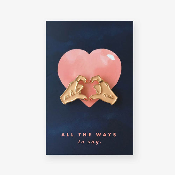 Hands of Love enamel pins by All the Ways to Say featuring gold-plated hands forming a heart with “u & me” text on pink and navy background.