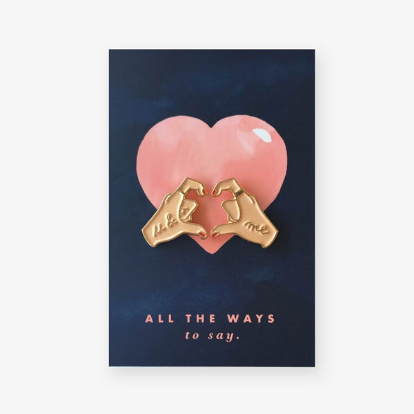 Hands of Love enamel pins by All the Ways to Say featuring gold-plated hands forming a heart with “u & me” text on pink and navy background.