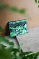 Green Cliff card holder displayed on a stone surface with tropical leaves in the background, elegant botanical pattern by All the Ways to Say.