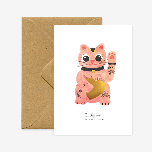 A greeting card featuring a illustration of a lucky cat with gold foil details, accompanied by a brown envelope.