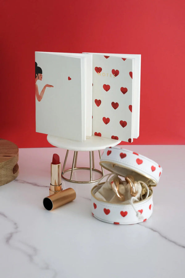 Romantic notebook with red heart design on white cover, displayed with jewelry case and lipstick against red background.