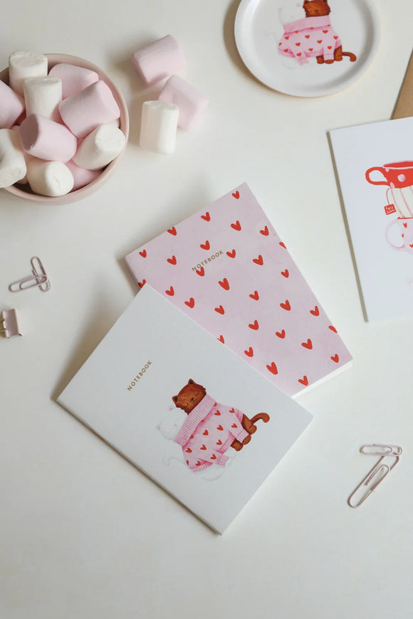 Set of cat-themed notebooks, one with illustrated cats in sweaters and one with red heart pattern, styled with marshmallows and stationery.