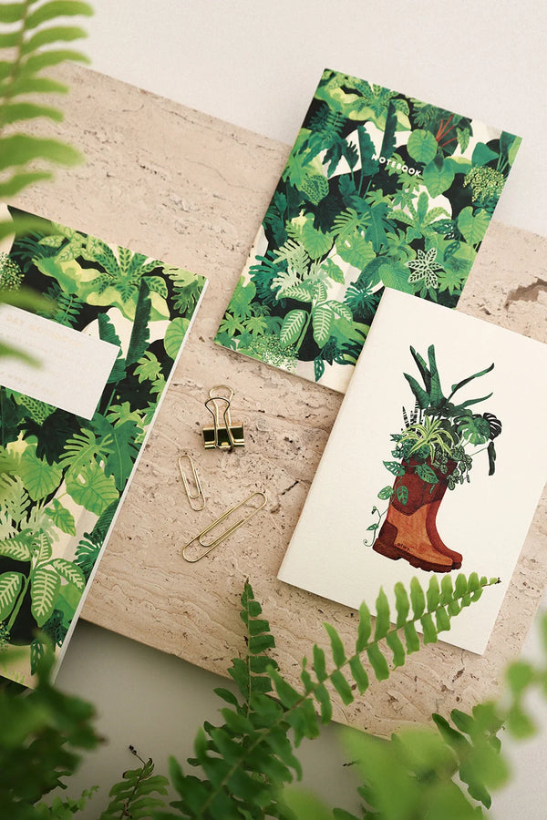 Botanical notebooks with green leafy designs and boot illustration, styled with paper clips and surrounded by fresh plants.