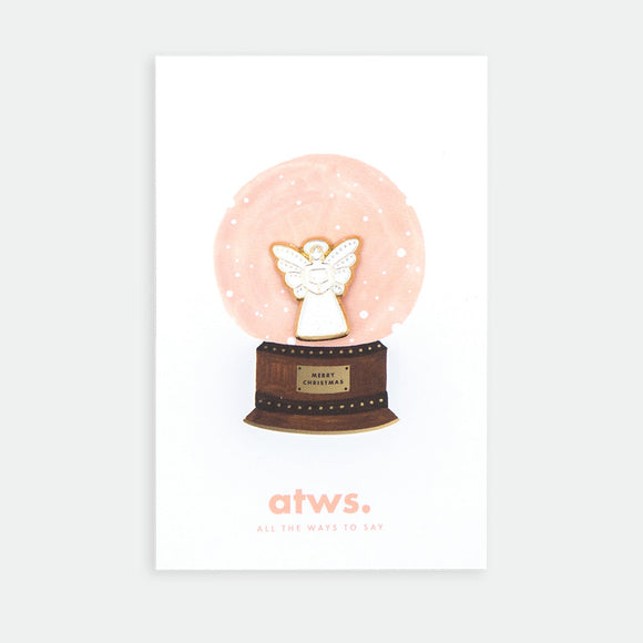 Angel enamel pin by All the Ways to Say featuring gold-plated angel design inside illustrated Christmas snow globe background with soft pink tones.