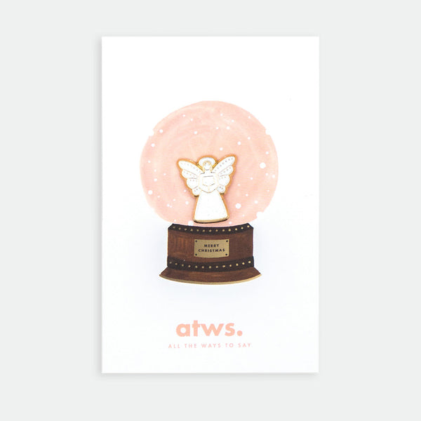 Angel enamel pin by All the Ways to Say featuring gold-plated angel design inside illustrated Christmas snow globe background with soft pink tones.