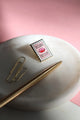 Bisou Bella enamel pin photographed on white marble tray with gold pen – artistic lighting and soft pink background