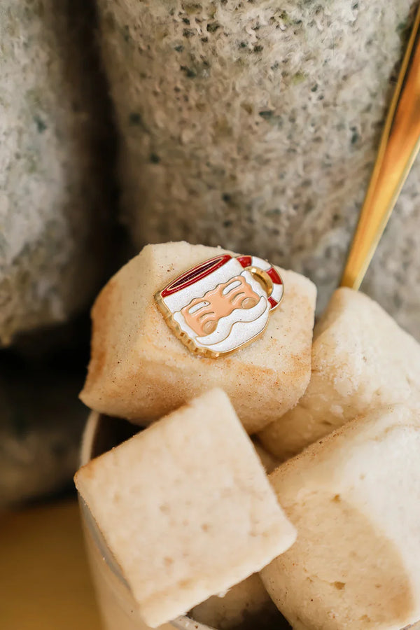 Santa’s Cup enamel pin by All The Ways To Say held in hands around a mug of marshmallows – soft and comforting winter vibe