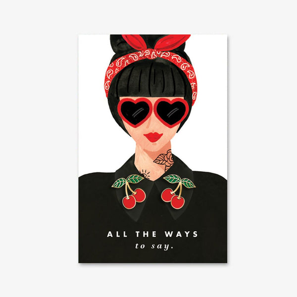 Cherries enamel pins set by All the Ways to Say featuring two gold-plated red cherries on illustrated retro woman background with heart-shaped glasses.