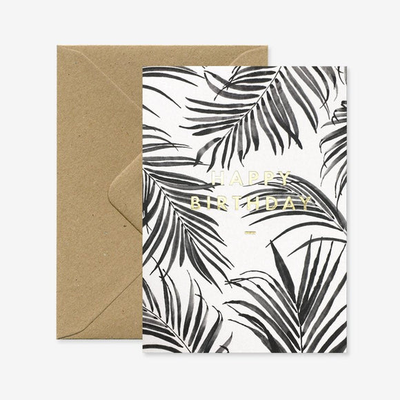 A birthday greeting card with a black and white palm leaf design and gold foil details, accompanied by a brown envelope.