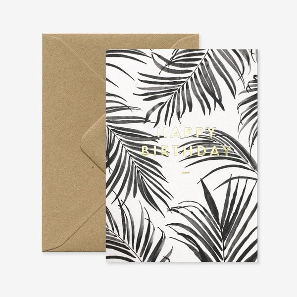 A birthday greeting card with a black and white palm leaf design and gold foil details, accompanied by a brown envelope.