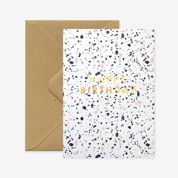 A birthday greeting card with a gold foil design and a speckled pattern, alongside a brown envelope.