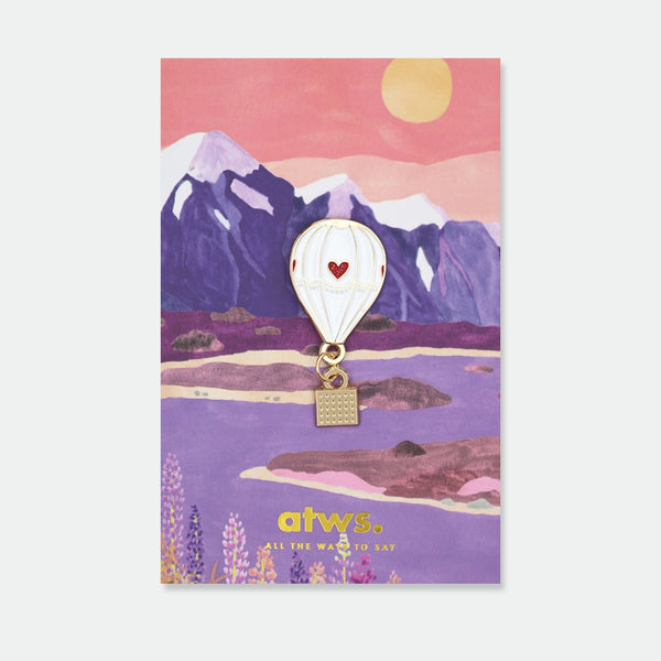 Enamel pin of a white hot air balloon with a glitter red heart, a small gold basket hanging below on a chain