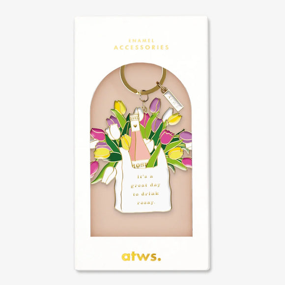 A stylish gold enamel keychain by All The Ways To Say featuring a white bag with a rosé bottle and tulips, inscribed with "it's a great day to drink rosay."