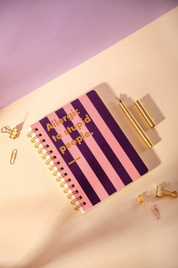 "Allergic to stupid people" A5 spiral notebook with pink and purple stripes, gold lettering and gold binding, next to a gold pen