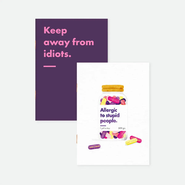 Allergic To Stupid People A6 pocket notebook duo covers by All The Ways To Say, one dark purple notebook with "Keep away from idiots." text in pink lettering, the other featuring an illustrated pill jar labeled "Allergic to stupid people, 1 pill a day, 200 gr."