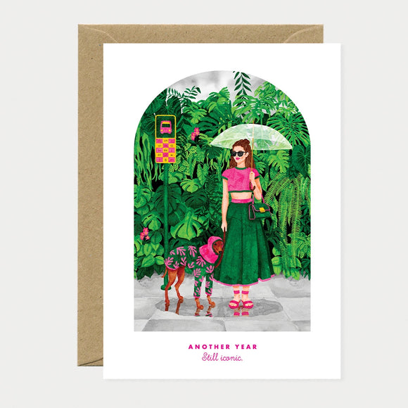 Illustrated birthday greeting card of a woman in a pink top and green skirt holding an umbrella, walking a dachshund dressed in a matching pink and green outfit, surrounded by lush greenery, text reading "Another year, still iconic"