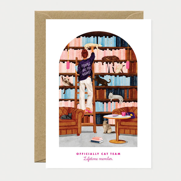 Illustrated greeting card of a woman on a ladder in a library, wearing a sweater that reads "People are the worst," surrounded by seven cats on the bookshelves, sofas, and floor, text reading "Officially cat team, Lifetime member"