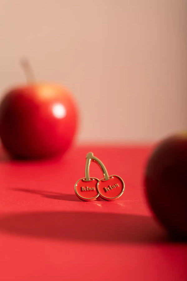 Kiss Kiss - Pins: artistic product photography of the cherry enamel pin on a red background with apples.