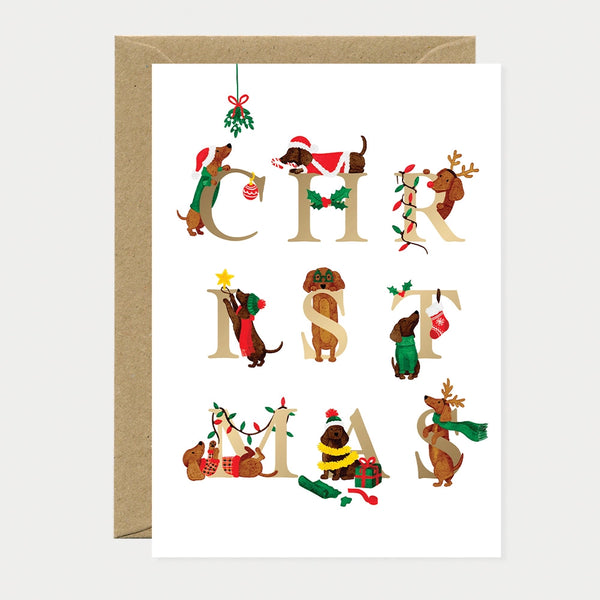 Illustrated Christmas card featuring nine dachshunds dressed in holiday outfits (Santa hats, reindeer antlers, scarves, sweaters) each posed with a gold foil letter spelling "CHRISTMAS"