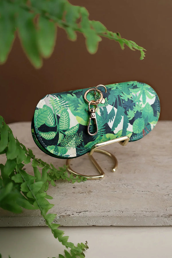 Green Cliff sunglasses case with tropical leaves print, gold clasp and keyring, displayed on marble stand with fern background.