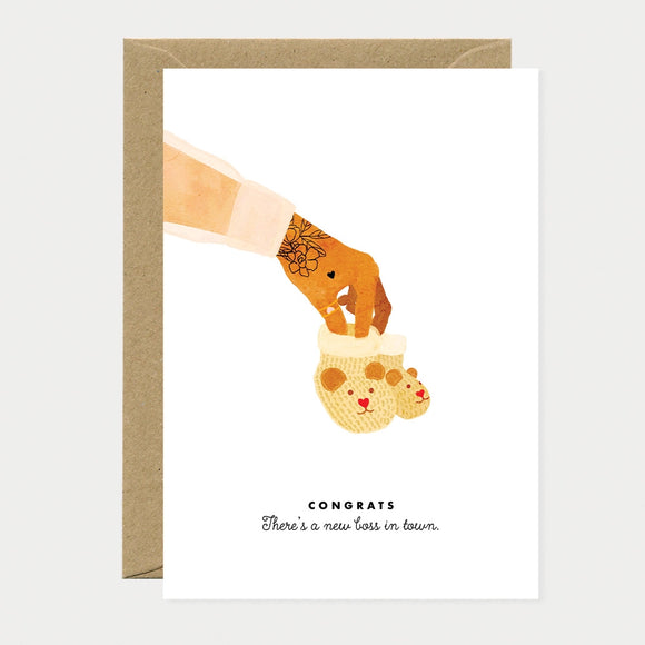 Illustrated greeting card of a tattooed hand with a finger inside a pair of yellow teddy bear-shaped baby mittens, text reading "Congrats, There's a new boss in town"
