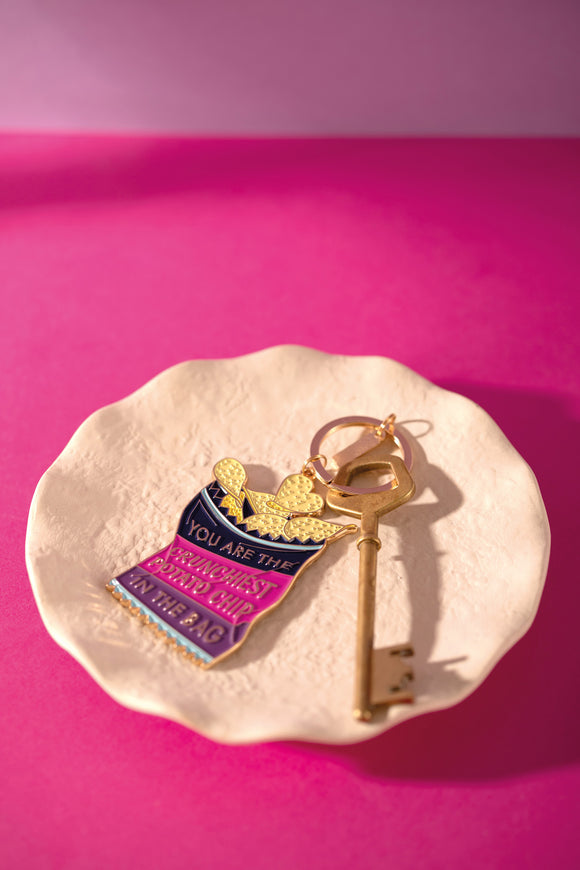 Potato chip bag enamel keychain reading "You are the crunchiest potato chip in the bag", with a gold key on a ceramic dish