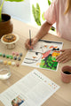 Dog Sitter paint by numbers in progress on a table with 15 paint pots, a glass of water, coffee and the color guide