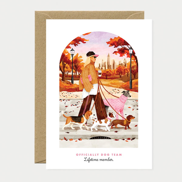 Illustrated greeting card of a woman walking four dogs through an autumn park with a city skyline in the background, text reading "Officially dog team, Lifetime member"