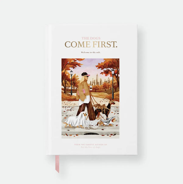 Dogs Come First hardcover A5 notebook by All The Ways To Say, cream cover styled like a book jacket with the title "The Dogs Come First, welcome to the cult," a fake author credit, and an illustration of a woman walking four dogs in Halloween costumes through an autumn park