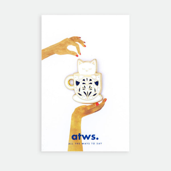 Cup Cat enamel pin by All the Ways to Say featuring gold-plated cat sleeping in teacup with blue pattern on illustrated hands background.