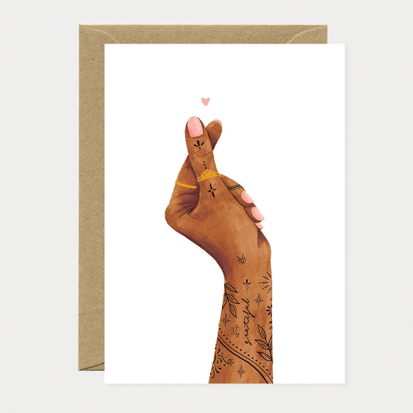 Illustrated greeting card of a tattooed hand forming a small heart shape with two fingers, a tiny pink heart floating above, the word "grateful" written along the forearm
